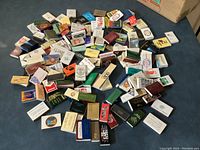 View showing a large assortment of approximately 100 empty matchboxes in varied colors and designs spread out on a blue surface.