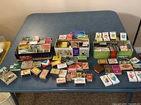 Collection of various matchbooks and matchboxes arranged in three metal and plastic boxes as well as spread on a table.