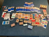 Wide view of collection with 52 empty matchboxes and matchbooks laid out on a blue surface, showing various colorful and vintage Swedish designs.