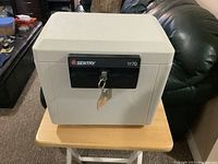 Front view of Sentry Safe Model 1170 showing light gray metal construction, black lock panel with key inserted, and two small compartments.