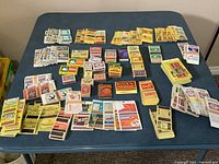Wide view of a large assortment of vintage matchbook covers and matchboxes of various brands and colors laid out on a blue table.
