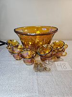 Photo showing large punch bowl surrounded by eight matching carnival glass cups and small accessories in plastic bag placed in front, all on textured light-colored surface.