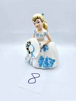 Front view of vintage porcelain girl vase holding a wide-brimmed hat with blue flowers and ribbon, showcasing detailed dress and painted features.