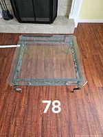 Square glass coffee table showcasing the clear glass top and decorative cast iron frame with scrolling design on hardwood flooring.