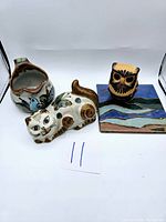 Full view of three pottery items: cat figurine, jug, and tile with owl. Background is white to highlight items.