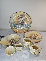 Overview of seven-piece Royal Doulton Famous Ships ceramic collection arranged on table, showing plates, bowl, pitchers, creamer, and sugar bowl with maritime imagery.