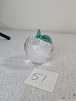 Clear glass apple-shaped paperweight with green glass leaf on top sitting on textured fabric with paper marked '51'
