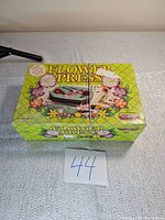 Top and side view of vintage flower press box showing colorful floral graphics and brand Little Genie.