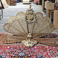 Front view of antique brass fireplace fan showing full spread of fan blades resting on decorative rug and behind it various furniture.