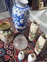 Photo showing all seven vases together on a patterned rug, including various shapes and colors.