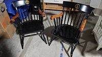 Two black spindle back Captain chairs with natural wood colored armrests and engraved emblem on top rail sitting side by side.