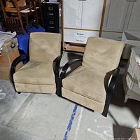 Two large beige upholstered contemporary accent chairs with dark curved wooden arms shown from front with chairs side by side on concrete floor.