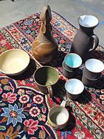 Overview of all ceramic serving pieces displayed on a patterned rug including pitcher, vases, cups, and bowl