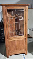 Full view of the mission style wooden corner cabinet with glass door showing leaded glass design on the door and wood paneling below.