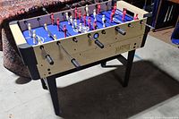 Full view of the Matrix gaming table showing foosball rods and player figures in red and white, blue playing surface, black legs and reinforced black corners.