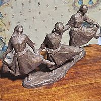 Front angled view of metal sculpture showing three dancing women holding hands.