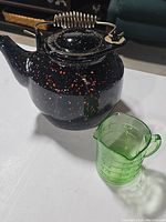 Tea kettle in black enamel with red and white speckles alongside green uranium glass measuring cup on a white surface