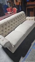 Front and left side view of the beige tufted sofa showing tufted backrest and arms, nailhead trim, and three seat cushions.