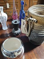Photo showing group of six vases including white ceramic vase with rose, pink glass vase, blue glass bottle, dark carved vase, clear crystal vase, and woven basket vase.