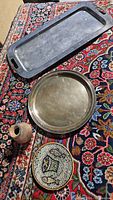 Four vintage items displayed on a patterned rug including a black rectangular tray, round metallic tray, small pottery vase, and mosaic plate.