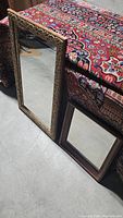Photo showing two mirrors side by side, one large with detailed carved wooden frame and one smaller with plain dark wood frame, both resting against a patterned rug and concrete floor.