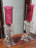 Full view of both vintage ruby etched hurricane lamps showing ruby red glass chimneys, clear glass bases with etched flowers and hanging crystal prisms.