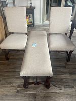 Front view of two upholstered dining chairs and an upholstered bench, all with light beige fabric and dark brown wooden frames.