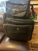 Photo showing the smaller computer shoulder bag on top of the larger rolling computer bag, both black. The shoulder bag has a logo and text embroidered in yellow and green on the front flap.