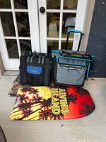 Photo shows Titan soft cooler with handle and wheels to the right, black and blue Lounge Lizard Plus beach seat on left, and Alvin's Island boogie board in front, yellow-orange design with palm trees.