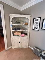 Full view of the wood corner cabinet painted white with distressed finish, placed in a corner with clothing items on open shelves and closed cabinet below.
