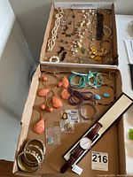 Top view of two cardboard trays with a mix of costume jewelry arranged inside, showing necklaces, bracelets, earrings, pins, and the Daniel Wellington watch.