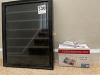 Frontal view of black framed glass display case with closed glass door and visible side clasp, cardboard box of 6 deck rotating card tray beside it