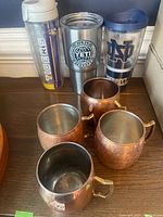 Front view of Yeti Rambler cup, two Tervis cups with sports logos, and four copper mugs with brass handles.