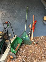 Green Scotts AccuGreen 3000 spreader and assorted lawn care tools leaning against wall with leaves on ground