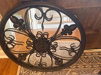 Photo showing round mirror with black metal frame and fleur de lis design, face up on patterned rug.