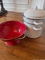 White enamel-coated pasta pot with lid and perforated insert shown next to a red colander.