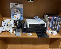 Shelf displaying Sony PS2 console with 4 controllers (3 Pelican white wired, 1 Chameleon in packaging), several PS2 game cases, Nintendo Wii remote sleeves and adapters.