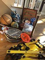 Standing assortment of sporting items on floor: basketball, helmets, rackets, footballs, glove, stuffed head, magnet board, chalk container.