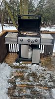 Outdoor black and silver gas BBQ grill with three control knobs, propane tank in base below grill, and two side metal shelves. Grill is on a wooden deck with some snow on ground.
