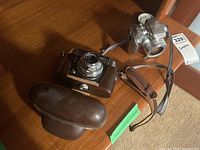 Two vintage cameras displayed, one Voigtländer in leather case and one Kodak camera.