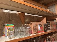 Shelf with multiple clear glass canning jars and lids, some jars inside cardboard boxes including Bernardin brand boxes.