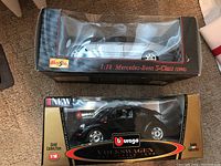 Two packaged model cars, a silver Mercedes Benz S-Class and a black Volkswagen Beetle, shown side by side on carpet surface.