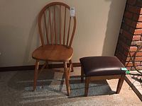 Photo displaying an oak wooden chair with a curved spindle back and a small oak stool with a dark brown faux leather cushioned seat, positioned on carpet near a brick structure.