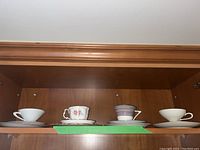 Three china tea sets displayed on a wooden shelf: one white with simple design, one with floral red gold design, one with pastel stripes.