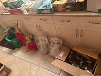 Photo showing Christmas decorations including gold deer figurines with red bows, plastic wrapped angel figurines, a red Christmas stocking, floral picks, and a glass jar arranged on a floor near cabinetry