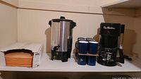 Wide shot showing Tim Hortons coffee maker, West Bend coffee percolator, twelve blue coffee mugs stacked in two rows, and a box of paper.