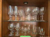 Two shelves of assorted glassware including crystal stemware and rocks glasses in a wooden cabinet.
