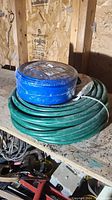 Two coiled hoses on a wooden shelf in an outdoor shed: blue 1.5 inch backwash hose wrapped in packaging tape and a green vinyl/rubber garden hose tied with a plastic strap.
