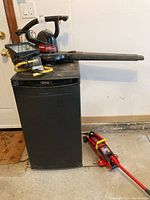 Toro Super Blower Vac sitting on top of a black Danby mini-fridge, with red Big Red hydraulic floor jack to the right side on concrete floor. Yellow cord coiled on work light on top of fridge.