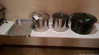 Four metal cooking vessels including two stainless steel pots with lids, one black enamel canning pot with lid, and one metal loaf pan arranged on a white shelf.